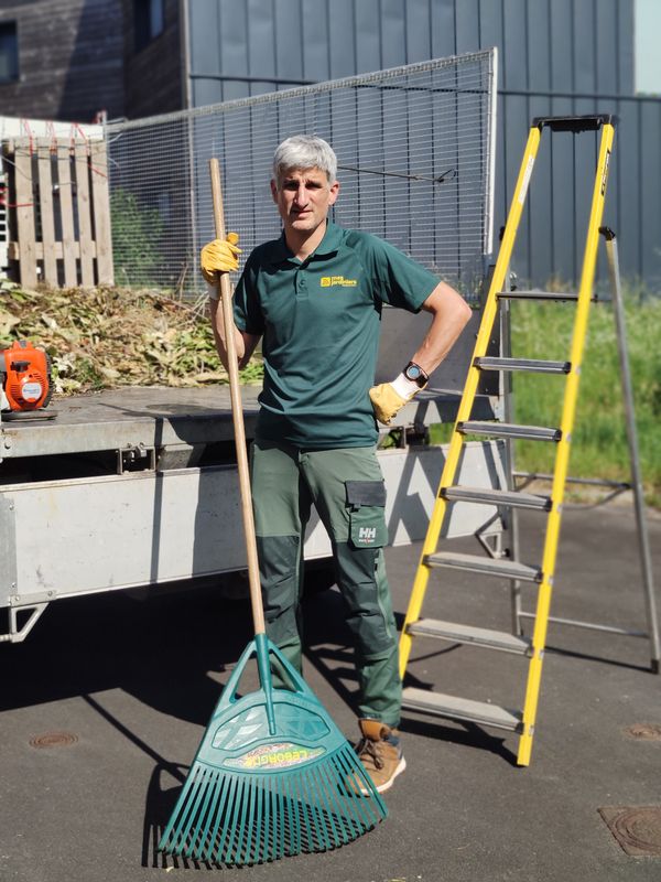 Jardinier Mes Jardiniers et son balai à gazon devant la benne chargée de déchets verts